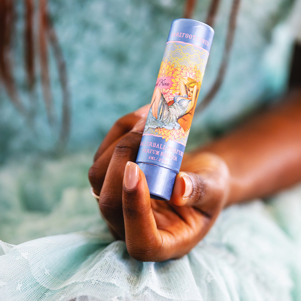 Person holding Barefoot Venus Coconut Kiss perfume oil with a woman in a blue dress and a flower background design being held by a woman in a blue dress.