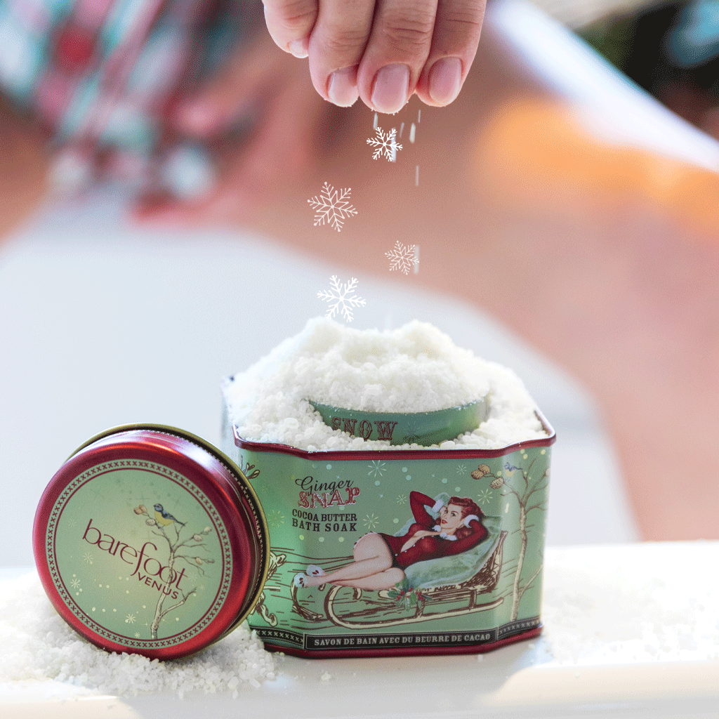 Tin of Barefoot Venus Ginger Snap bath soak filled with snow featuring a woman in a red outfit laying in a toboggan and a green background design on the packaging and animated snowflakes falling into the tin.