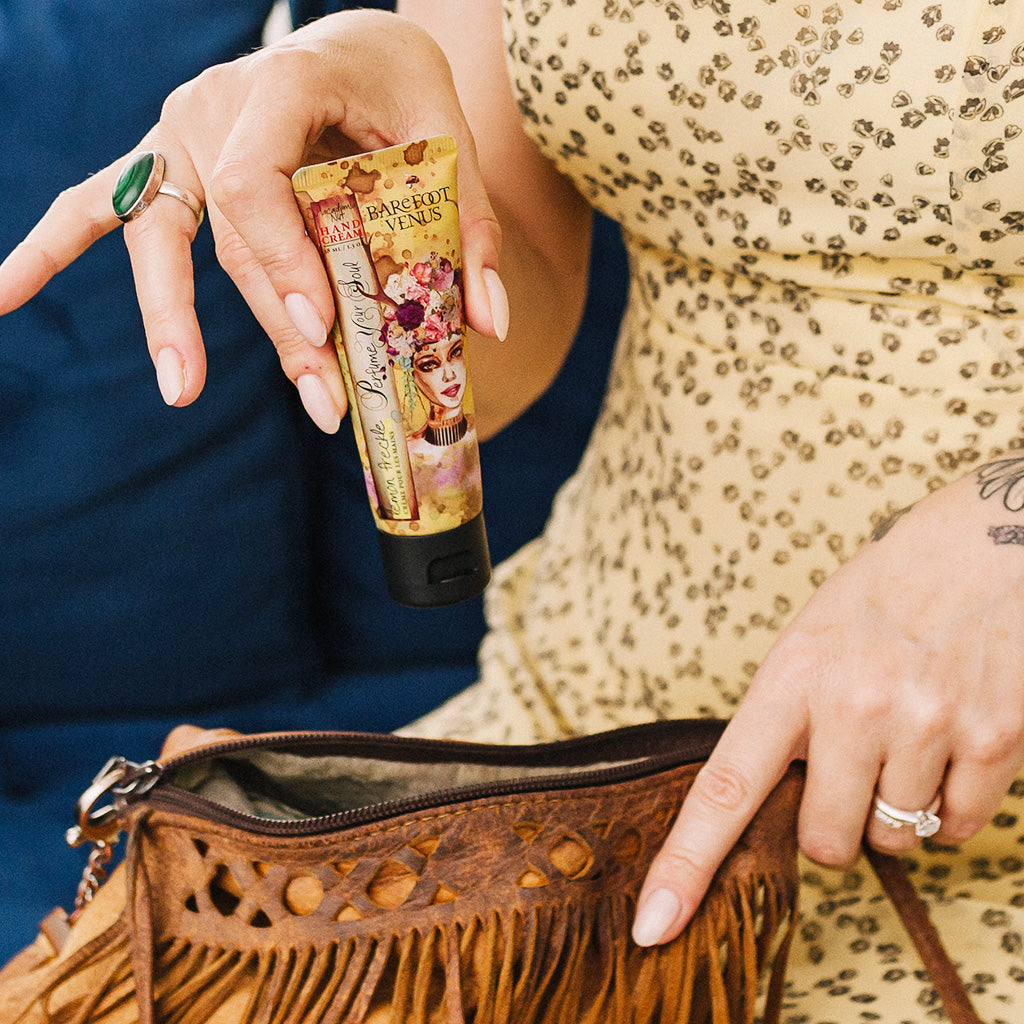 Woman holding Barefoot Venus lemon freckle hand cream above a fringe purse, while wearing a yellow dress.