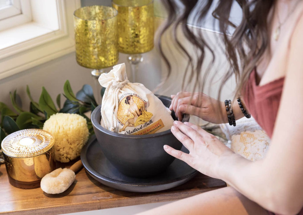 Person admiring canvas bag of Barefoot Venus mustard bath, bath soak in a large bowl with items surrounding it.