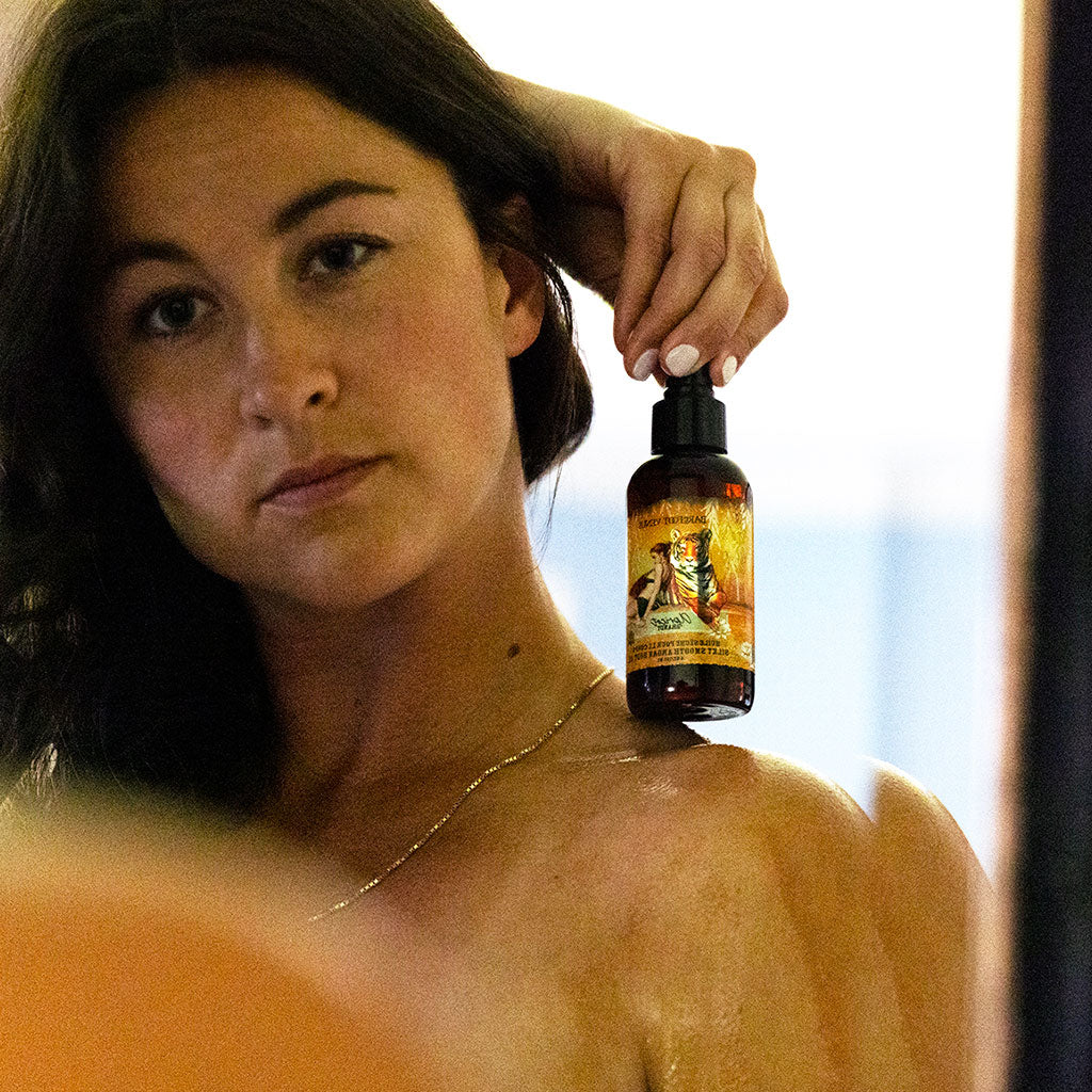 A spray bottle of Barefoot Venus Apricot Brandy Argan body oil with a black lid and a design of a tiger and a woman in a green outfit on a white background.
