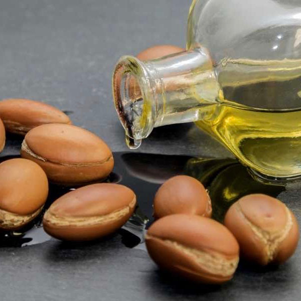 A clear glass bottle pouring out Argan body oil, alongside several Argan nuts on a dark background.