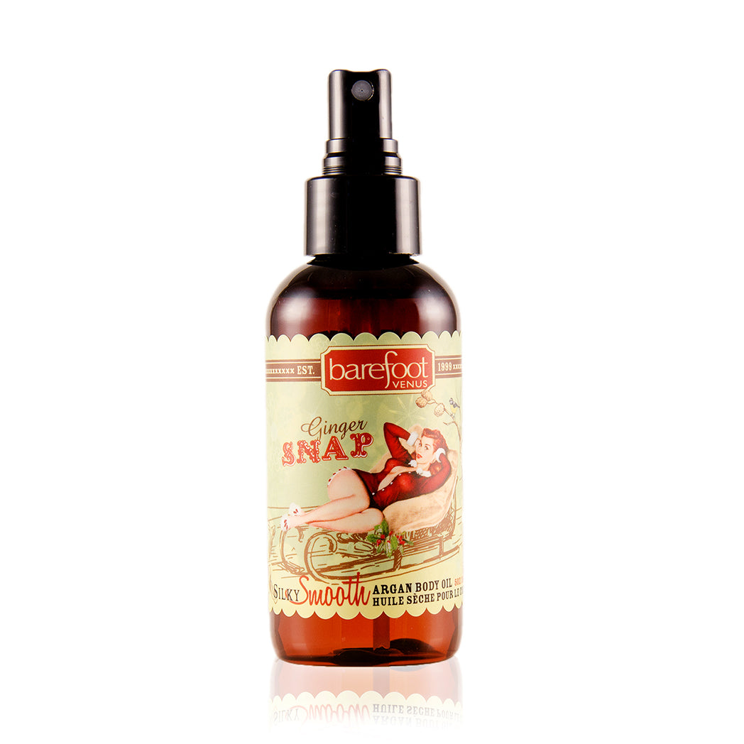 A spray bottle of Barefoot Venus Ginger Snap Argan body oil with a woman in a red outfit lying in a toboggan with a green background design on the packaging on a white background.
