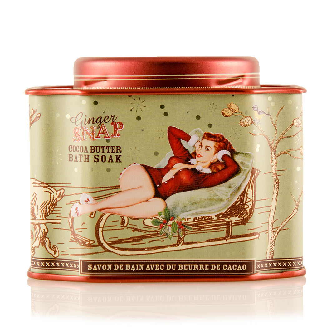 Tin of Barefoot Venus Ginger Snap bath soak with a woman in a red outfit lying in a toboggan with a green background design on the packaging on a white background.