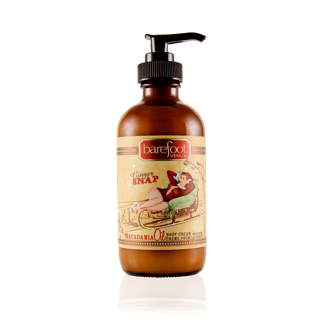 A dark brown bottle with a pump dispenser of Barefoot Venus Ginger Snap macadamia oil body cream product featuring a woman in a red outfit lying in a toboggan design on a white background.