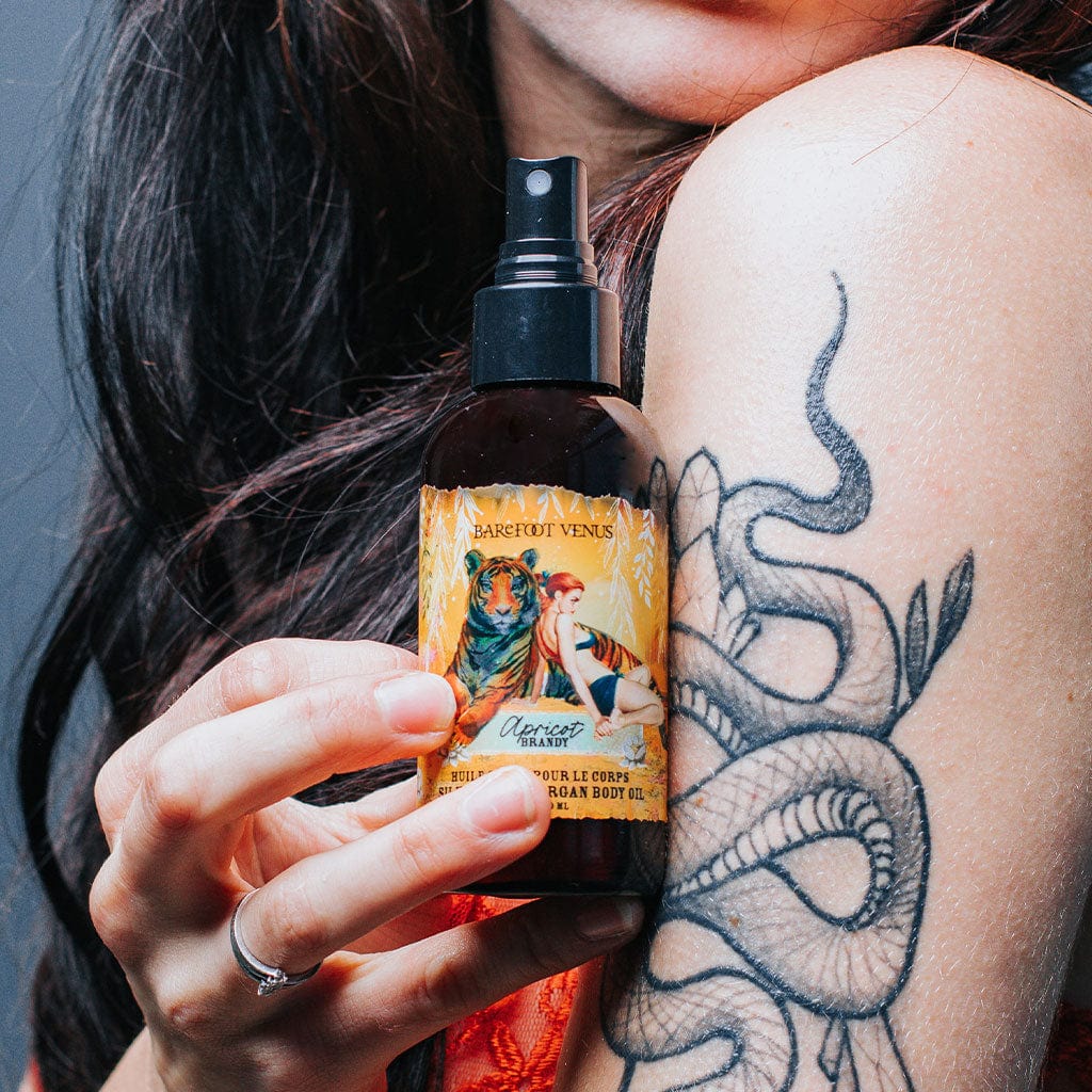A person holding a spray bottle of Apricot Brandy argan body oil with a label featuring illustrations of tiger and woman.