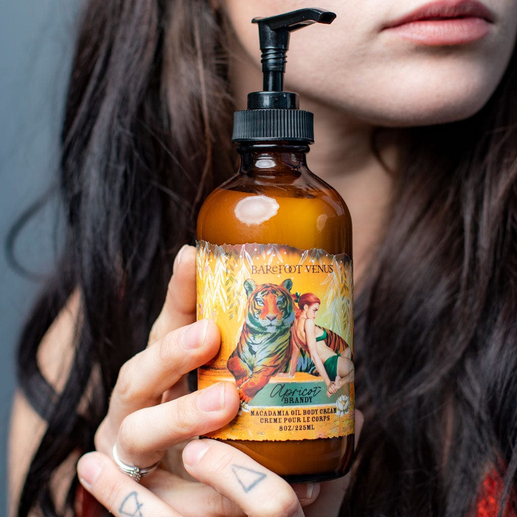 A person holding a pump bottle of Apricot Brandy body cream with a label featuring illustrations of tiger and woman.