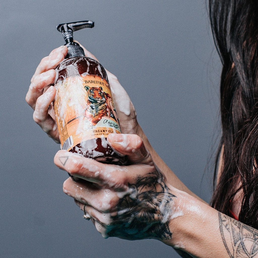 A person holding a pump bottle of Apricot Brandy creamy cleansing wash covered in bubbles with a label featuring illustrations of a tiger and woman.