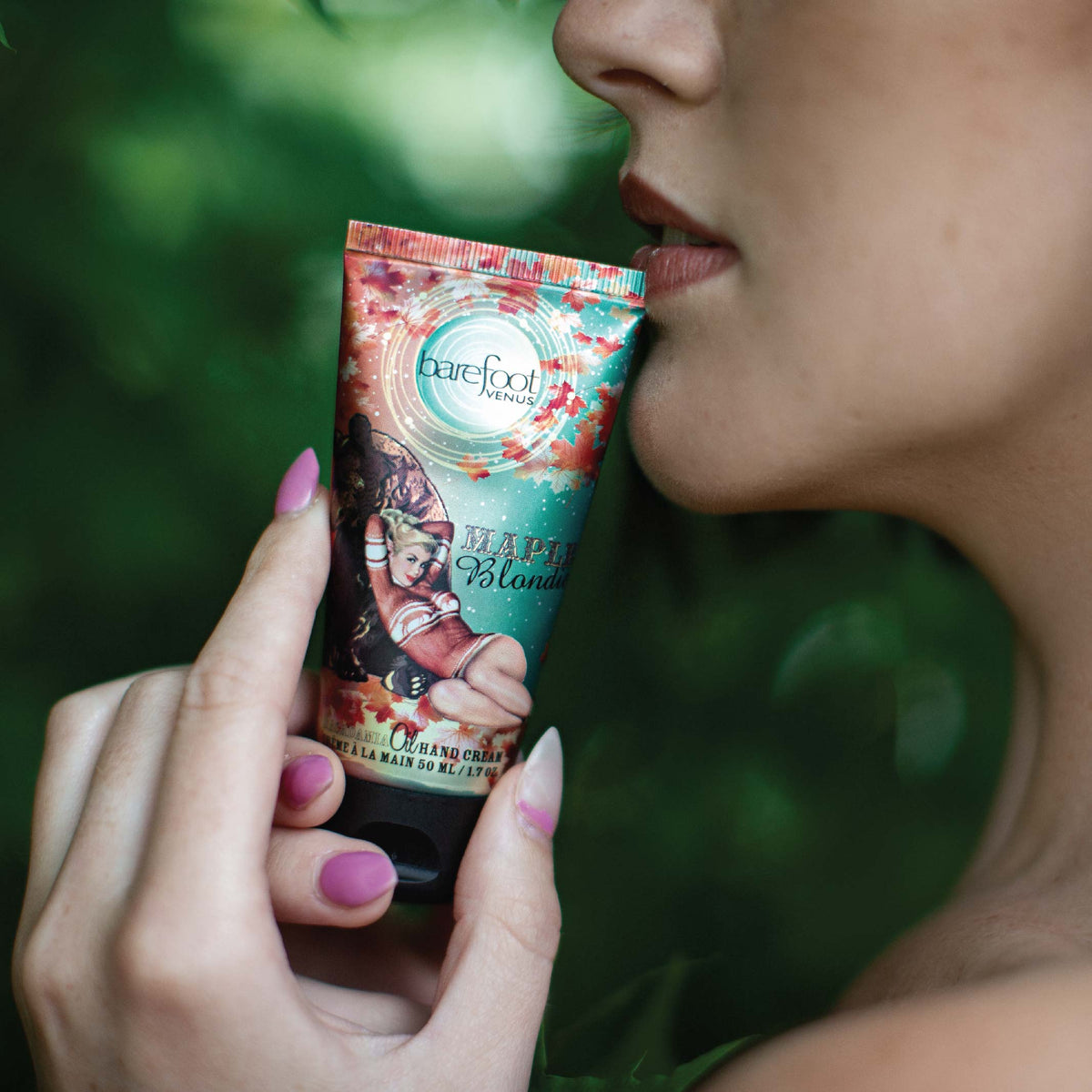 Person holding Barefoot Venus Maple Blondie hand cream with decorative packaging of bear and woman in a red sweater against chin.