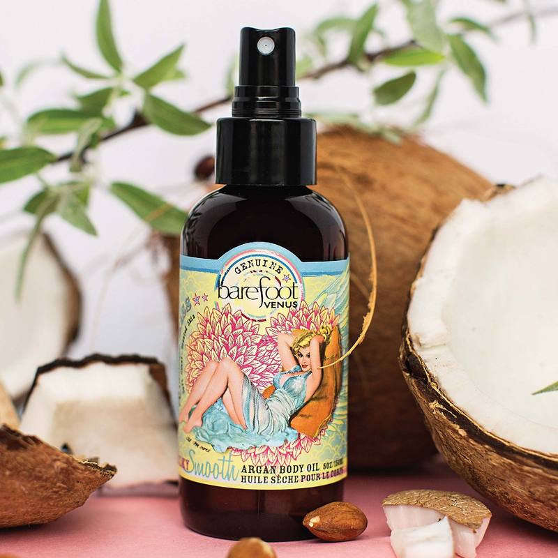 Spray bottle of Barefoot Venus Coconut Kiss Argan body oil product featuring a woman in a blue dress with a floral background design on a background of coconuts.