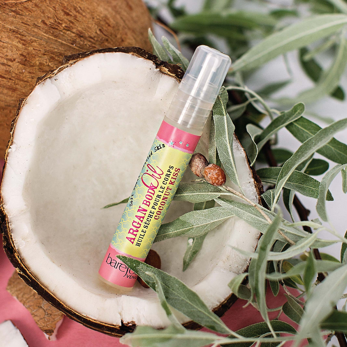 Spray bottle of Barefoot Venus Coconut Kiss Argan body oil mini product inside a coconut with greenery in the background.
