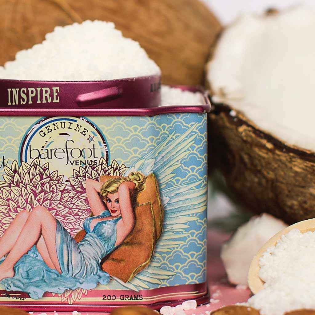 Tin of Barefoot Venus Coconut Kiss bath soak product featuring a woman in a blue dress with a floral background design with a background of coconuts.