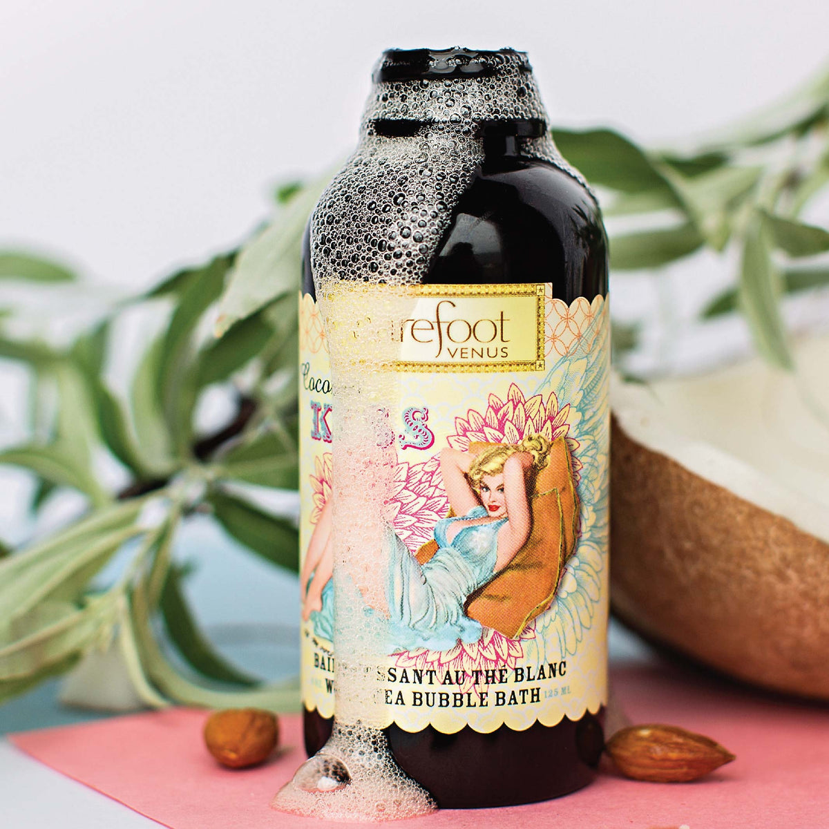 Bottle of Barefoot Venus Coconut Kiss bubble bath product featuring a woman in a blue dress with a floral background design with a blurred background.