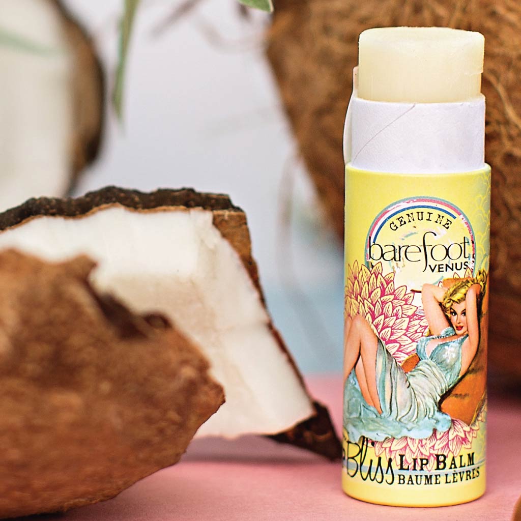 Tube of Barefoot Venus Coconut Kiss lip bliss featuring a woman in a blue dress with a floral background design with a background of coconuts.