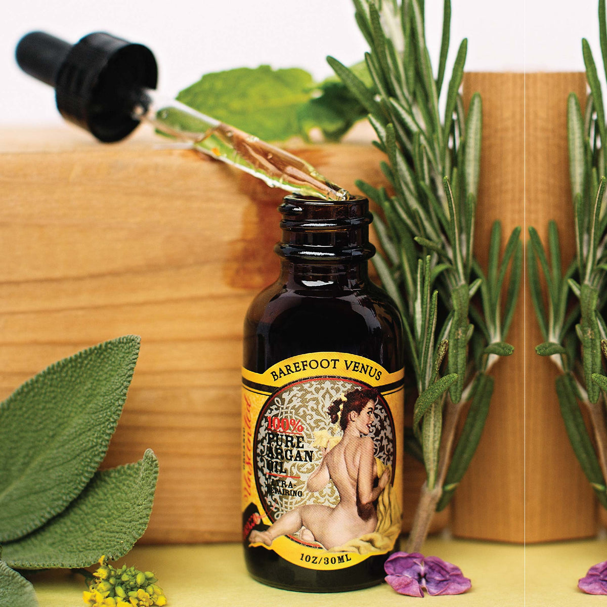 A dropper bottle of Barefoot Venus 100% Pure Argan oil with a vintage design on the packaging with a wooden greenery background.
