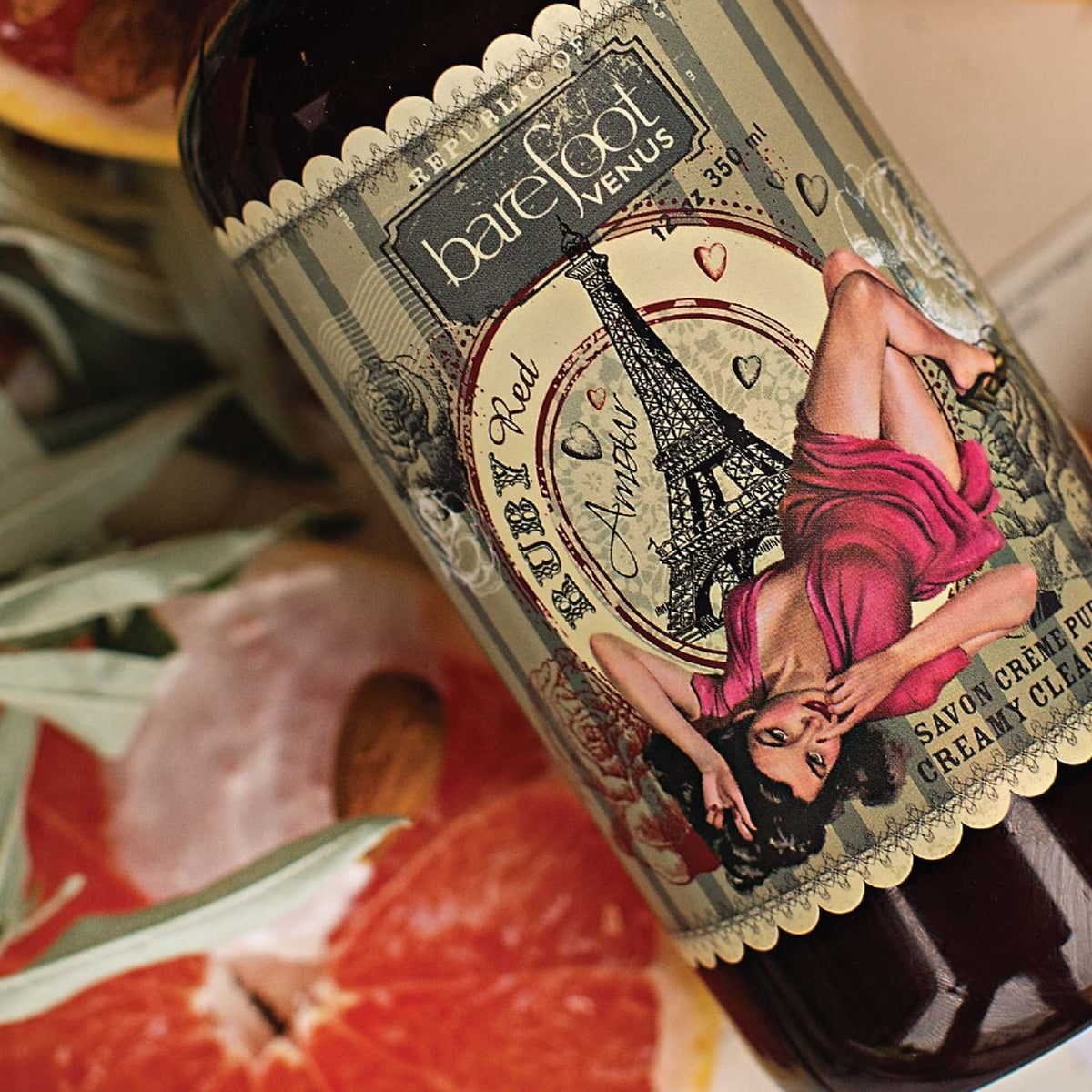 Close up of a bottle of Barefoot Venus Ruby Red creamy cleansing wash product featuring a woman in a red dress in front of the Eiffel tower with background of grapefruits, almonds and leaves.