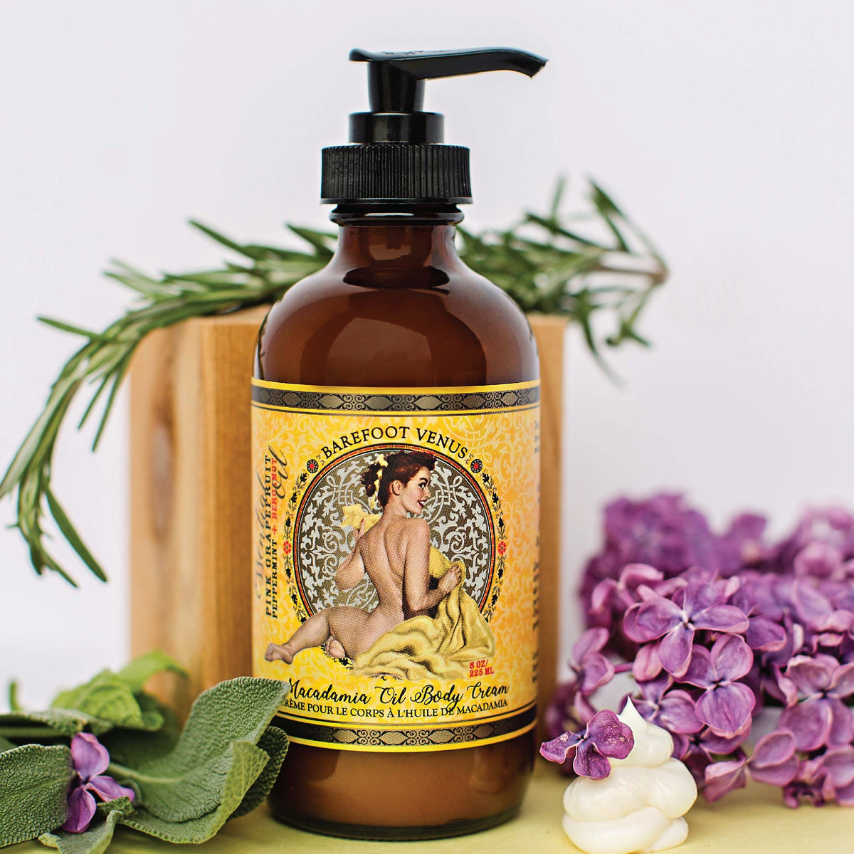 A dark brown bottle with a pump dispenser of Barefoot Venus Coconut Kiss Macadamia Oil body cream with flowers and greenery in the background.