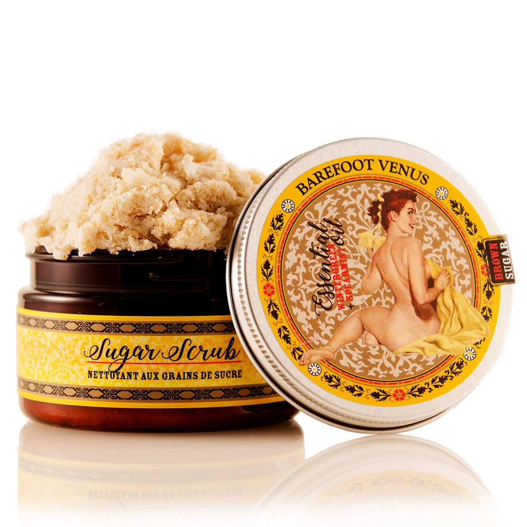 Overflowing container of Barefoot Venus Essential Oil sugar scrub with the lid beside the product container with a vintage woman design on the packaging on a white background.