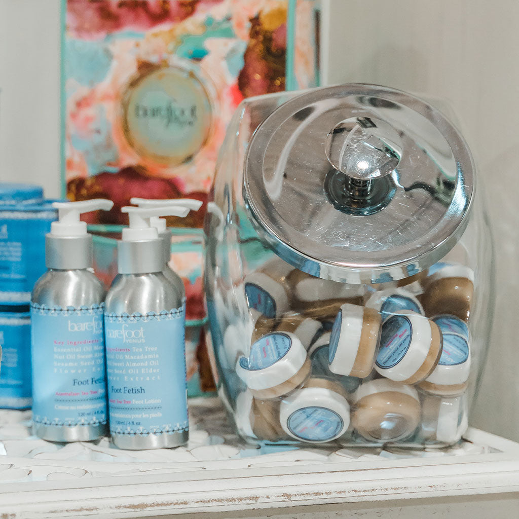 A display of Barefoot Venus foot care products including Foot Fetish lotions on a shelf, and All Cracked Up foot balm arranged in a clear glass container with metal lid, on a wooden shelf.