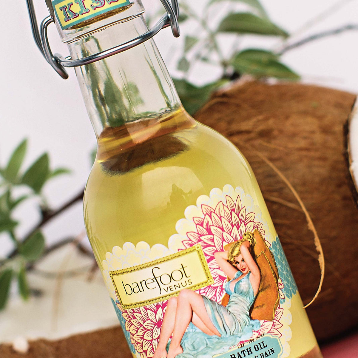 A close up of a bottle of Barefoot Venus Coconut Kiss massage & bath oil featuring a woman in a blue dress with a floral background design on packaging and a background with coconuts.